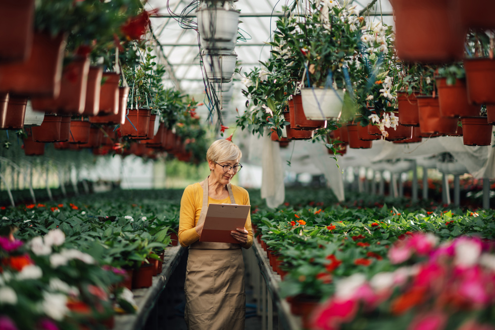 Portrait of senior female florist standing at greenhouse full of greenery, plants and blooming colorful flowers and writing in clipboard while tracking growth process. Old botanist checking flowers